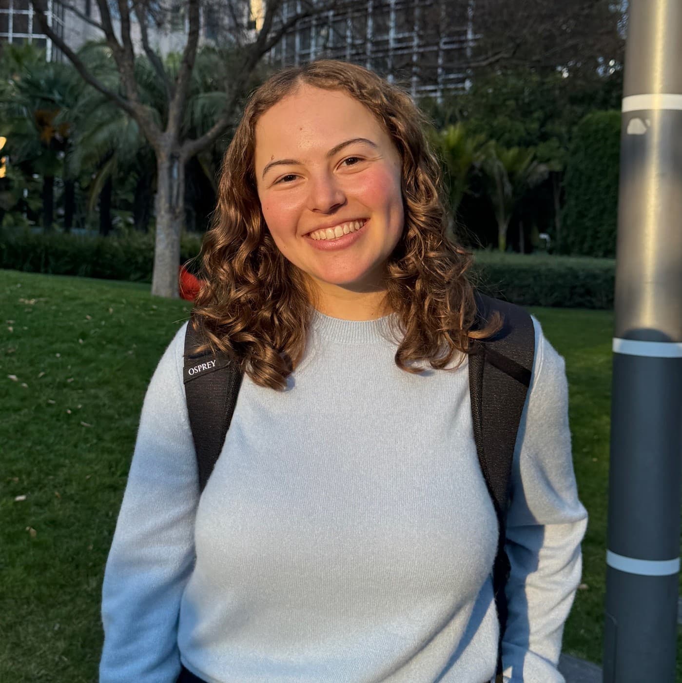 Becca Pollard standing on a lawn in a city park, wearing a light blue jumper and a backpack, smiling at the camera.